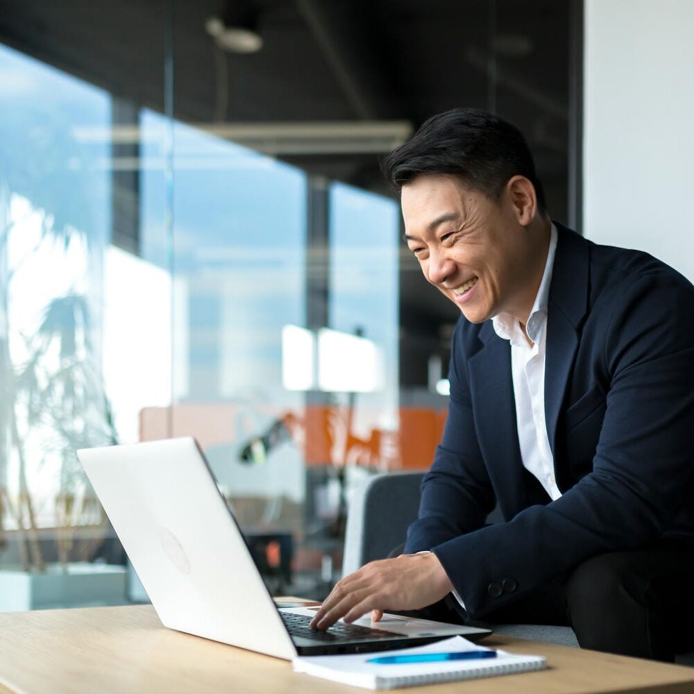 Senior Asian business man working on laptop online, smiling and rejoicing, happy boss business owner working in office