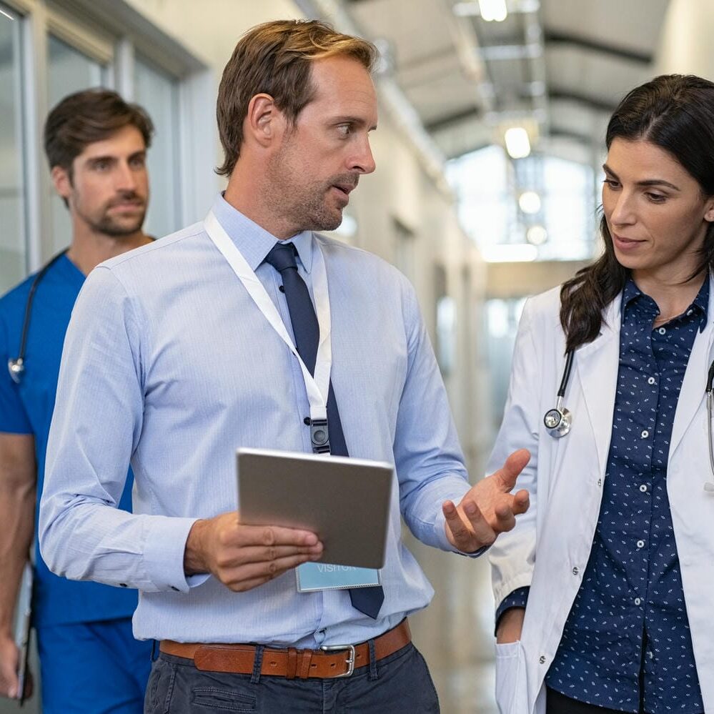 Man and woman doctor having a discussion in hospital hallway while holding digital tablet. Doctor discussing patient case status with his medical staff after operation. Pharmaceutical representative showing medical report.